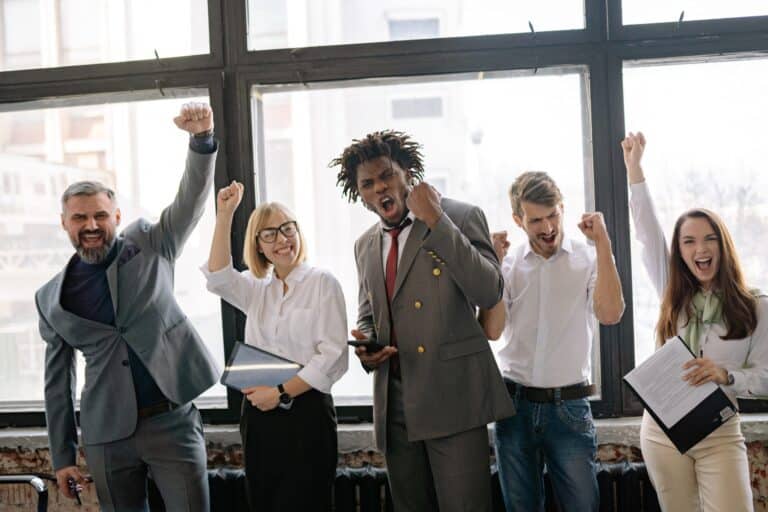 5 people in an office with smiles on their face and their hands raised cheering with happiness