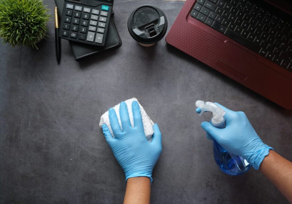 Hands with gloves, a towel, and cleaning spray wiping down a desk